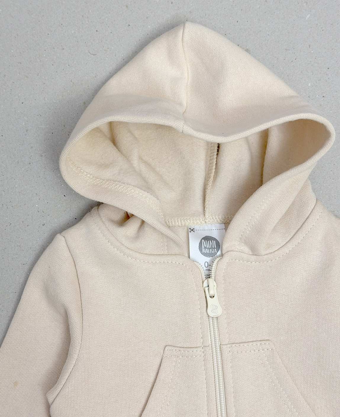 Casaco Moletom Bege MiniMalista — detalhe dos bolsos frontais funcionais que aquecem as mãozinhas e punhos em ribana canelada que bloqueiam entrada de vento. Anti-pilling premium mantém malha lisa lavagem após lavagem.